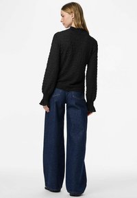 Crni teksturirani top s dugim rukavima i rubovima s volanima, uparen s visokim strukom i širokim plavim trapericama. Pogled sprijeda straga.