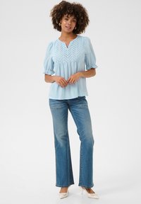 Világoskék, rövid ujjú felső V-nyakkal és díszes lyukasztott mintával; kiflibe szabott farmernadrággal és bézs magassarkúval párosítva. Sima textúra.