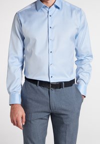 Ljusblå långärmad skjorta med nedknäppt krage, med en slät textur och glänsande knappar, ihop med grå strukturerade byxor och ett svart bälte.