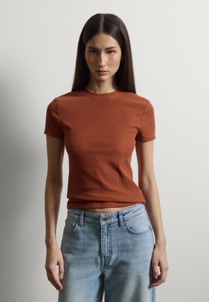 Junge Frau mit langen, dunklen Haaren, die ein kurzärmeliges Oberteil in Rostfarbe und hellblaue, hoch taillierte Jeans trägt, vor einem einfarbigen Hintergrund stehend.