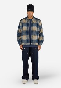 Plaid, blue and beige fleece jacket with a zip front, paired with dark blue wide-leg jeans and white sneakers. Black beanie worn on the head.
