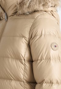 Beige vadderad jacka med huva i päls och en cirkulär logotyp på överarmen, framdragkedjan synlig.