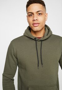 Olijfgroene hoodie van zachte stof met een capuchon met een trekkoord, voorzien van zwart-witte gemêleerde koorden en een frontale kangoeroezak.