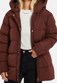 Femme portant une doudoune bordeaux avec une capuche, un haut blanc côtelé, un jean noir et un collier chaîne en or, les mains près des poches.