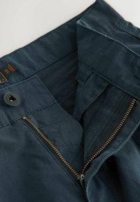 Pantalon bleu marine avec une fermeture éclair en laiton partiellement dézippée et un bouton triangulaire noir sur la ceinture portant la taille 37R.