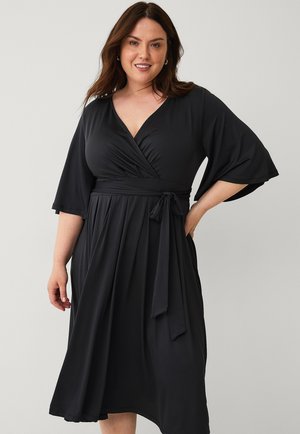 Zwarte wikkeljurk gemaakt van zacht materiaal. Heeft een diepe V-hals, wijde mouwen, een strikceintuur rond de taille en een vloeiende zoom.