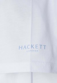 Camiseta de algodón blanca con el logo "HACKETT LONDON" en azul claro. Textura suave, corte regular, diseño limpio, de un solo color sin patrones.