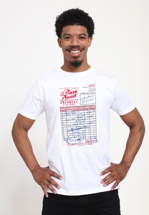 Lächelnder Mann mit lockigem Haar, der ein weißes T-Shirt trägt, auf dem eine rote und blaue Pizza Planet-Bestellquittung aus Toy Story abgebildet ist.