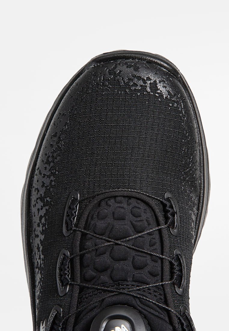 Chaussure de sport noire avec une tige en tissu texturé, un bout arrondi et des accents brillants. Détail notable : un système de laçage central.