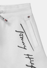 Weiße Sweatpants mit einem verstellbaren Bund, Seitentasche und schwarzem grafischem Textdesign. Der Stoff wirkt weich und hat eine glatte Textur.