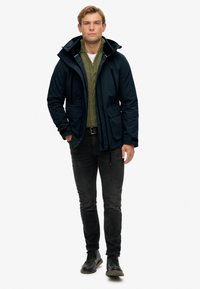 Chaqueta impermeable azul marino con capucha, dos bolsillos frontales, superpuesta a un suéter de punto verde, combinada con jeans negros y zapatos oscuros.