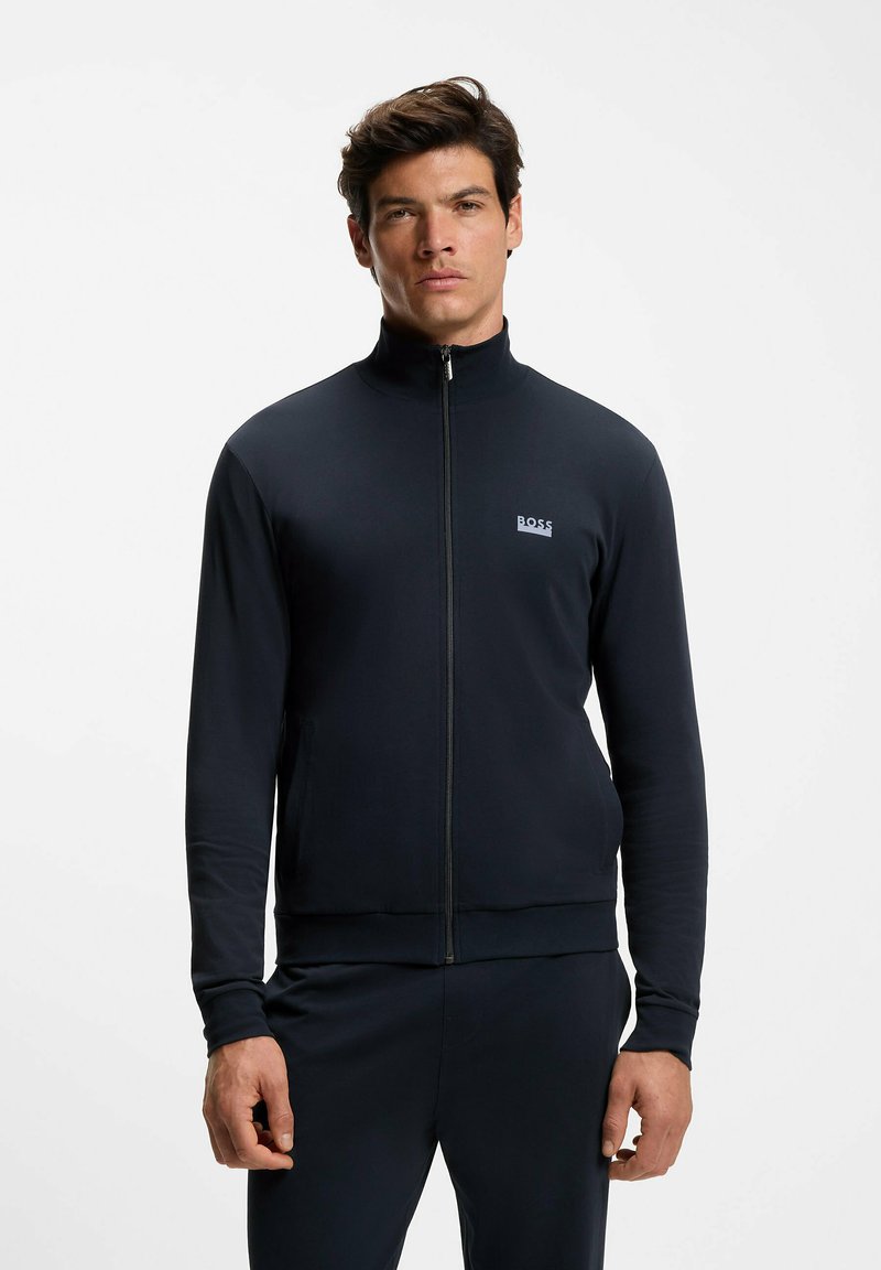 Laivastonsininen vetoketjullinen takki, valmistettu sileästä kankaasta, jossa korkea kaulus, resorit hihansuissa ja helmassa sekä pieni valkoinen logo rinnassa.