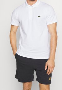 Polo-shirt blanc à manches courtes et col, avec un logo crocodile vert sur la poitrine gauche. Associé à un short foncé, offrant un look sportif.