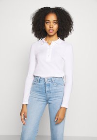 Vit långärmad polo-tröja med krage och plaggknäppning med tre knappar, i kombination med ljusblå högmidjade jeans. Slät textur, avslappnad passform.