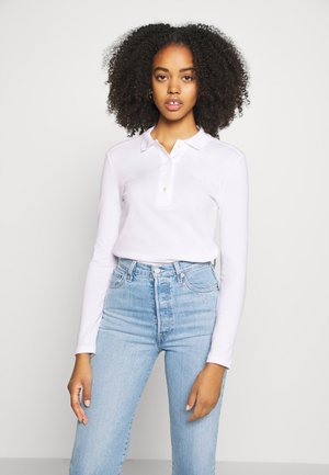 Maglietta polo bianca a maniche lunghe con colletto e placket a tre bottoni, abbinata a jeans chiari a vita alta. Tessuto morbido, vestibilità casual.