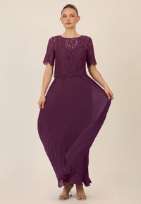 Pitkä violetti mekko pitsipäällä ja laskostetulla hameella, jossa on lyhyet hihat, pyöreä kaula-aukko sekä kellotettu, röyhelöillä varustettu helma.