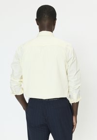 Chemise jaune pale à manches longues avec poignets boutonnés et col classique. Portée avec un pantalon à rayures bleu marine et une ceinture noire tissée.