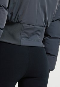 Giacca piumino grigio scuro con vita a costine, accentuazione del logo sul lato sinistro e texture liscia, abbinata a leggings neri.