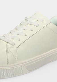 Gros plan d'une basket basse blanche avec des lacets blancs et une semelle en caoutchouc texturée sur un fond clair.