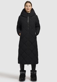 Lang sort quiltet frakke med hætte, der har en lynlåslukning fortil og sidelommer, parret med sorte ankelstøvler og tætsiddende sorte bukser.