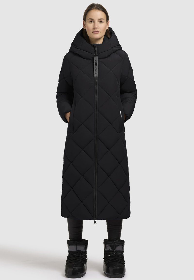 Lang sort quiltet frakke med hætte, der har en lynlåslukning fortil og sidelommer, parret med sorte ankelstøvler og tætsiddende sorte bukser.