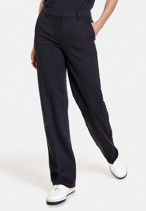 Person iført sorte bukser med lige ben og sidelommer samt hvide sneakers, stående med det ene ben let fremad.