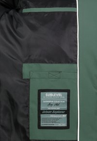 Grüne Oberbekleidung mit schwarzem Innenfutter, ausgestattet mit einer Tasche und einem Etikett mit der Aufschrift "SUBLEVEL" sowie Pflegeanweisungen und einem Logo.