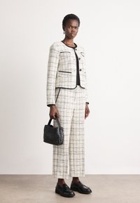 Costume en tweed à motifs en damier blanc et noir, avec une veste cropped et un pantalon à jambes larges. Accompagné d'un sac à main noir et de mocassins noirs.