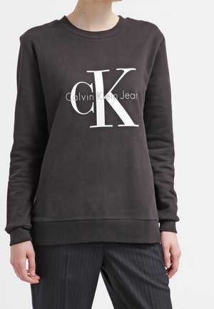 Osoba ubrana w czarną bluzę Calvin Klein Jeans z białym logo "cK" oraz czarne spodnie w prążki, stojąca na jednolitym tle.