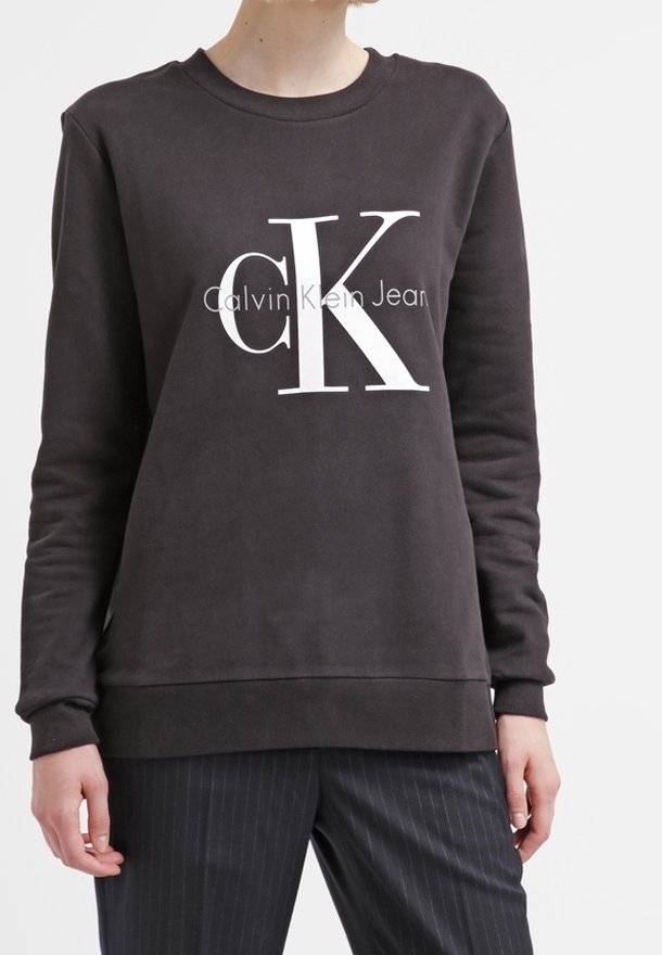 Personne portant un sweat-shirt noir Calvin Klein Jeans avec un logo "cK" blanc et un pantalon noir à fines rayures, debout devant un fond uni.
