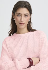 Pull en maille rose avec texture côtelée, doté d'un large décolleté. Un bracelet bordeaux orne le poignet, mettant en valeur le design de la tenue.