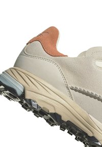 Beige mockasin sneakers med en tan accent vid hälen, texturerad mellansula och en svart greppyttersula. Har sömdekor.