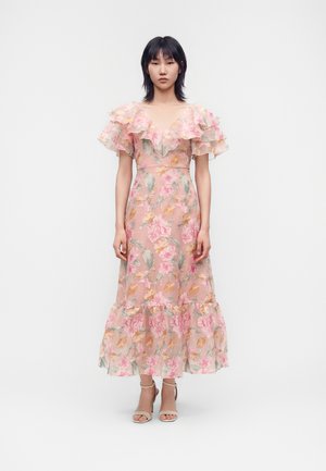 Femme portant une robe midi rose fleurie avec des manches et un ourlet à volants, debout devant un fond clair uni.