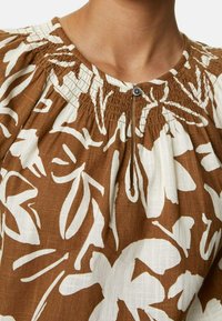 Blouse à motif floral marron et blanc avec un col froncé orné de smocks et une fermeture par bouton sous le col.