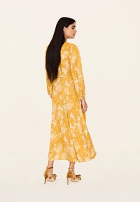 Rochie lungă galbenă cu flori, cu mâneci lungi și o siluetă fluidă. Prezintă o textură moale și modele florale intricate în alb și verde.