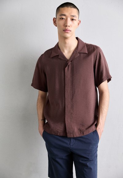 Chemise marron à manches courtes avec un fini texturé et un col, associée à un short bleu marine foncé, portée par une personne avec les mains dans les poches.