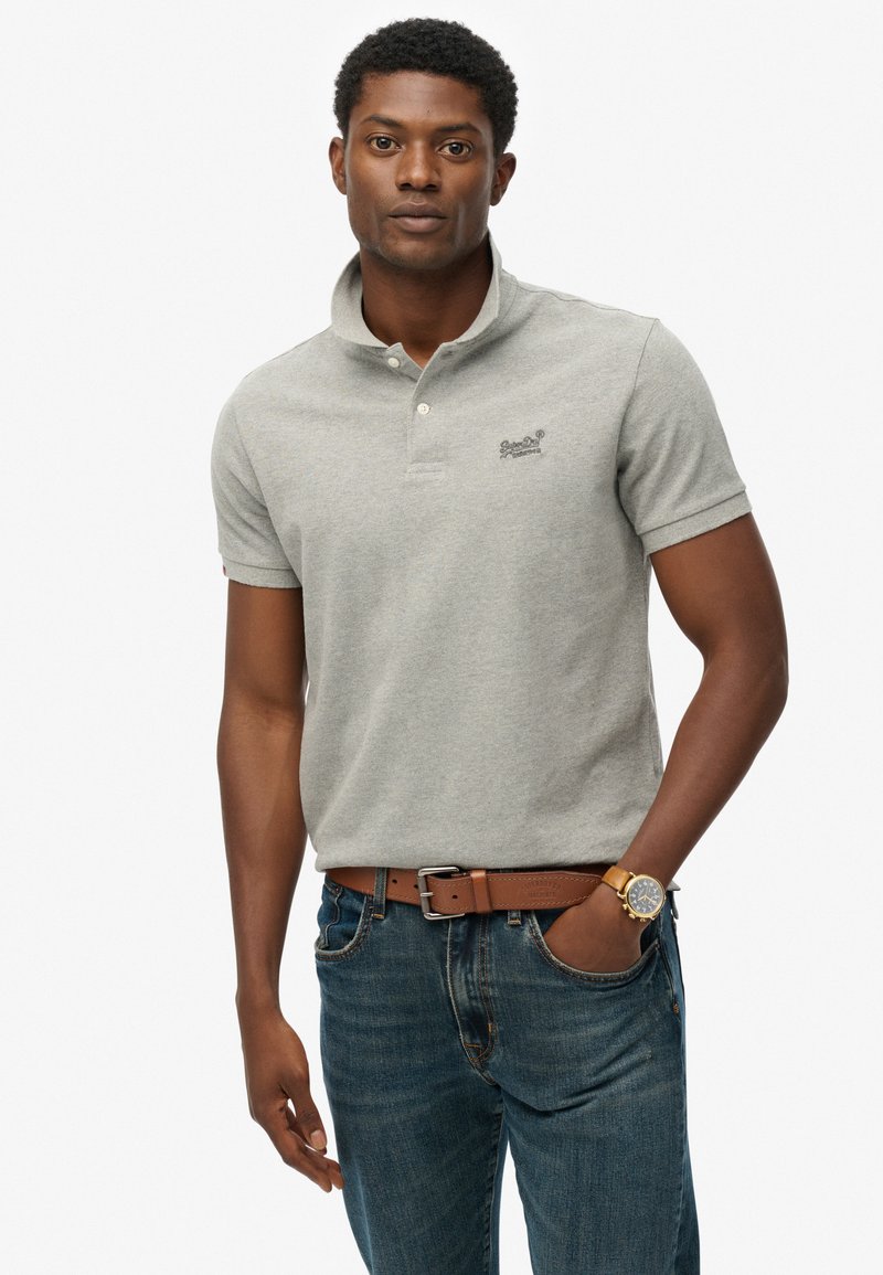 Polo gris en coton avec une patte de boutonnage à trois boutons et un logo brodé. Associé à un jean bleu et une ceinture marron, avec une montre.