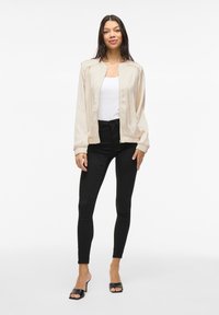 Beige bomberjacka i slät tyg, svarta slim-fit byxor och svarta öppna klackar. Jackan har elastiska muddar och en avslappnad passform.