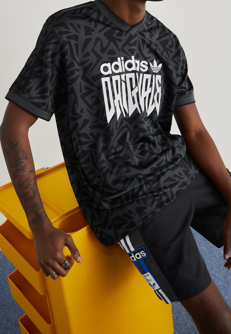 Černý adidas dres s krátkým rukávem, který má texturovaný grafický vzor a výrazný bílý text "ORIGINALS" na přední straně, s typickým výstřihem do V.