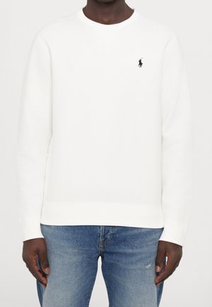 Osoba ubrana w prostą, białą bluzę z długim rękawem, z małym czarnym wyszywanym logo na lewej piersi, w połączeniu z niebieskimi dżinsami.