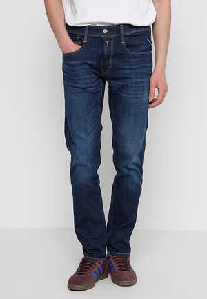 Dark blue denim jeans with a slim fit, classic five-pocket design, and contrasting yellow stitching. Worn with burgundy sneakers.