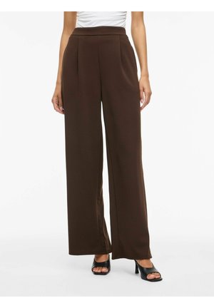 Femme portant un pantalon marron taille haute à jambes larges associé à des talons noirs ouverts, debout devant un fond blanc uni.