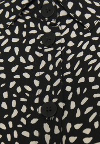 Μαύρο ύφασμα με ακανόνιστα λευκά στίγματα, με τρία μεγάλα μαύρα κουμπιά και μέρος γιακά.