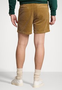 Man som bär bruna kordshorts, krämfärgade strumpor, vita sneakers och en mörkgrön långärmad tröja, står med ryggen mot kameran.