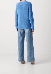 Woman wearing a bright blue knit sweater, light blue wide-leg jeans, and beige high-heeled shoes, standing against a plain wall.