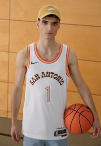 Camiseta de baloncesto blanca con acentos naranjas, que presenta "SAN ANTONIO" en texto marrón y el número "1" de un jugador. Sostiene un balón de baloncesto naranja. Gorra amarilla.