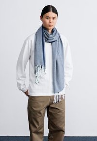 Blue scarf made of soft fabric, featuring a textured pattern and fringed ends, worn over a white long-sleeve shirt and beige pants.