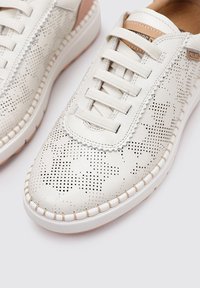 Zapatillas blancas de cuero con parte superior perforada, acentos en un tono marrón claro en contraste, cordones planos y suela texturizada. Detalle de costura elegante a lo largo de los bordes.
