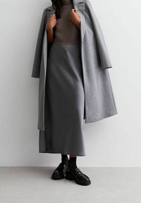Abrigo de lana gris sobre una blusa de canalé marrón claro, combinado con una falda larga gris y mocasines de charol negros. Texturas suaves y líneas limpias.