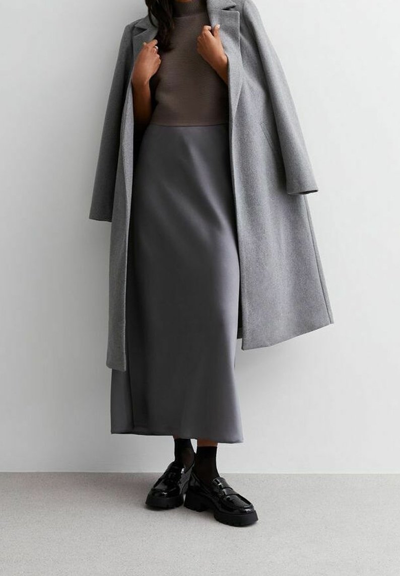 Abrigo de lana gris sobre una blusa de canalé marrón claro, combinado con una falda larga gris y mocasines de charol negros. Texturas suaves y líneas limpias.
